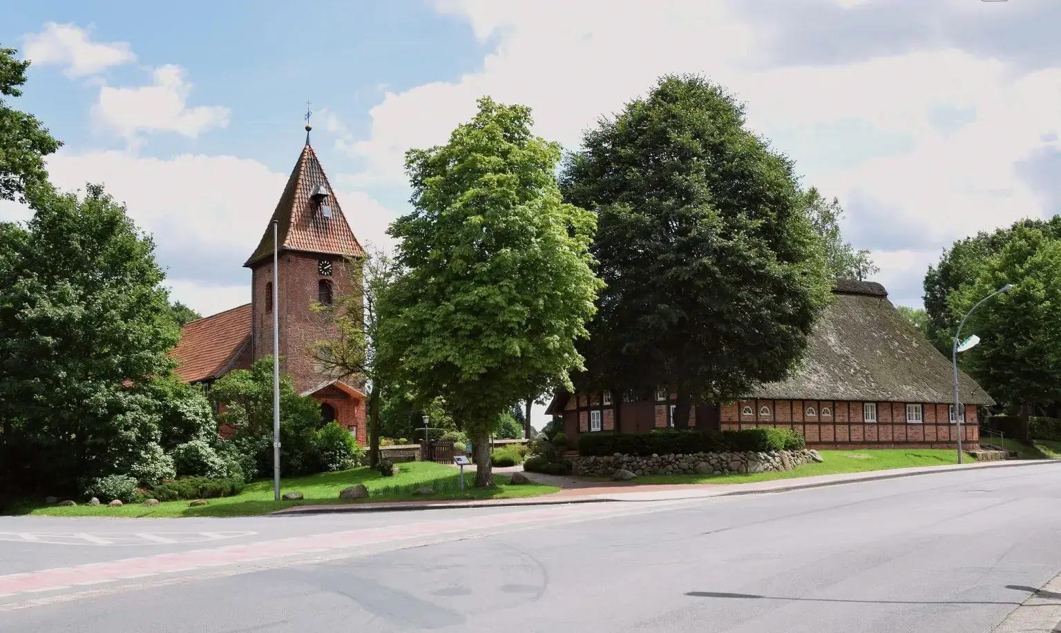 Historische Kirche und reetgedecktes Fachwerkhaus im Ortszentrum von Schwanewede, umgeben von Bäumen an einer ruhigen Straße.
