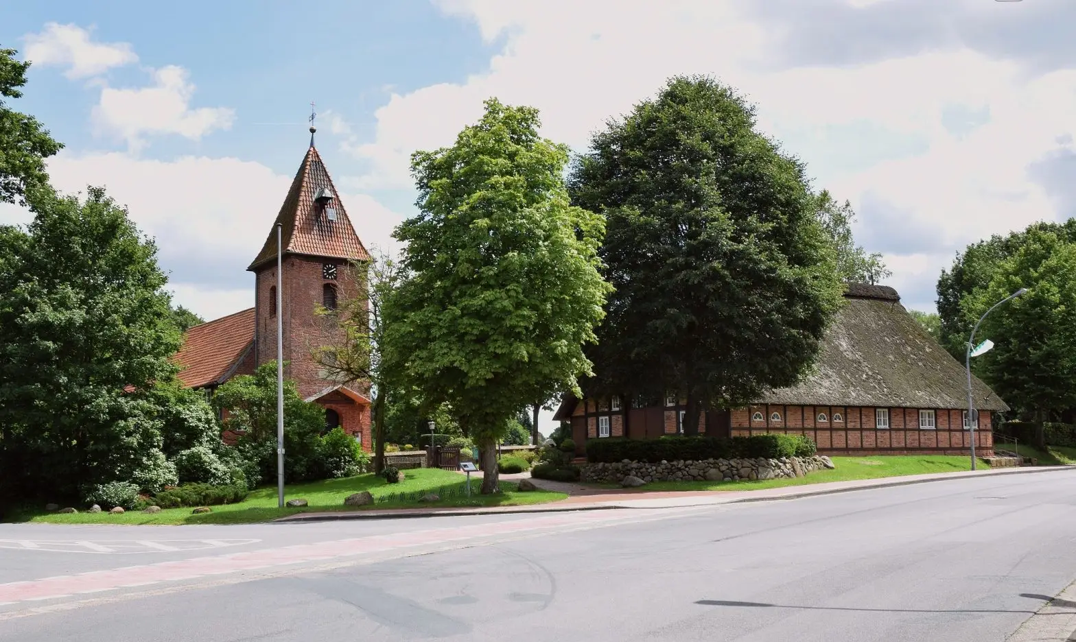 Historische Kirche und reetgedecktes Fachwerkhaus im Ortszentrum von Schwanewede, umgeben von Bäumen an einer ruhigen Straße.