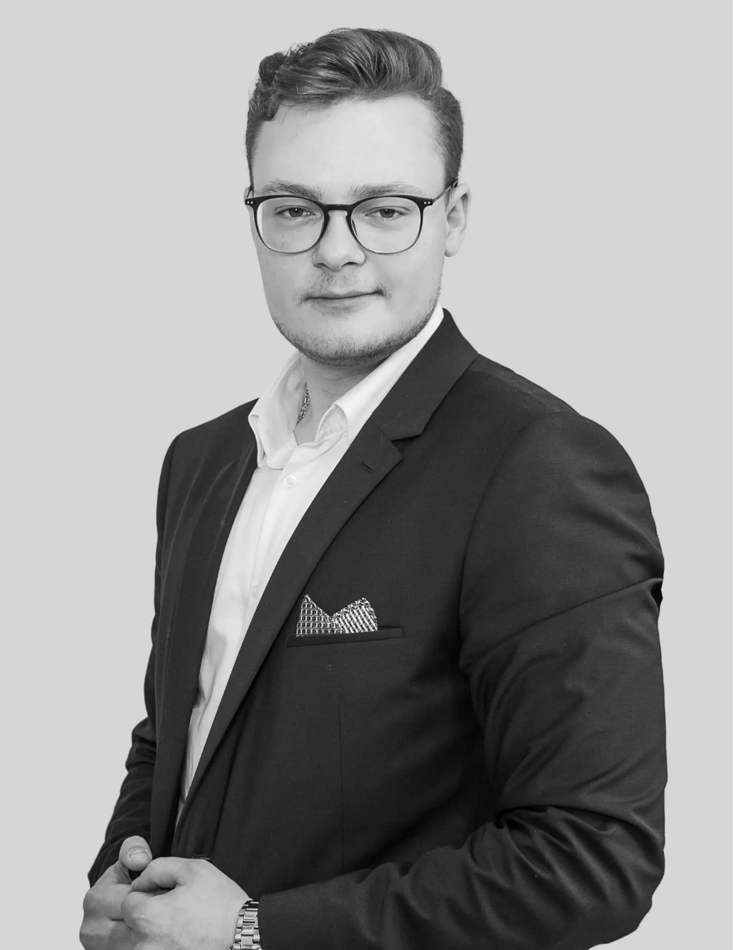 Porträtfoto eines jungen Mannes mit Brille in Business-Kleidung. Er trägt einen Anzug mit Einstecktuch und blickt mit leichtem Lächeln in die Kamera. Schwarz-weiß-Aufnahme vor neutralem Hintergrund.